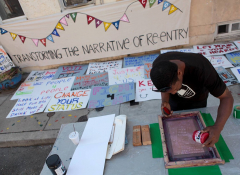 Image of an African American artist silk screening a poster with the text "Transforming the Narrative of Re-Entry" painted on a banner behind him. 