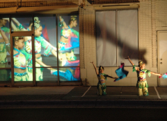 Evening, two dancers on a street with more dancers projected behind them.