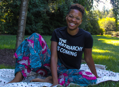 A young Black woman sitting on the grass wearing a shirt that says "The matriarchy is coming"