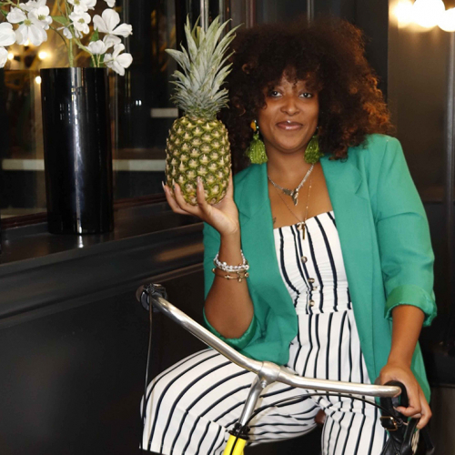 A young Black woman in a green blazer on a bicycle holding a pineapple. 
