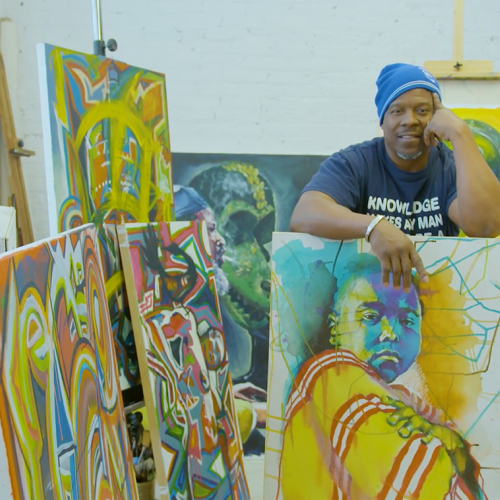 A black artist standing behind his paintings