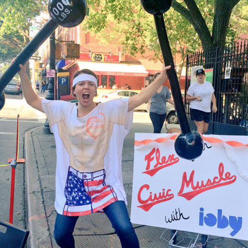 Image of a woman holding two plastic dumb bells with a tshirt that says “Civic-Muscle Tee."