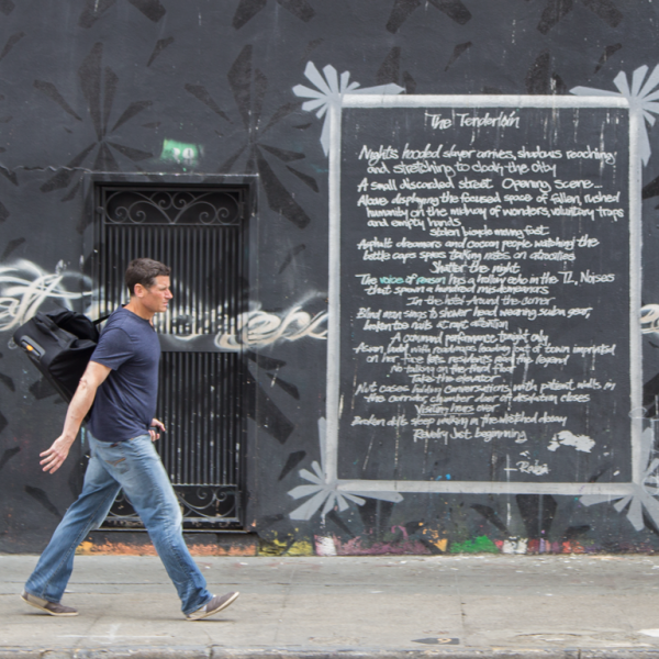 Image of a white man walking down the street in front of a piece of street art
