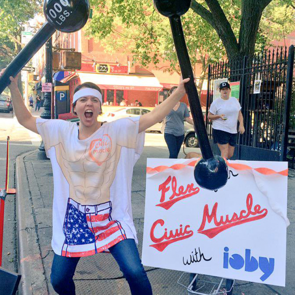 Image of a woman holding two plastic dumb bells with a tshirt that says “Civic-Muscle Tee."