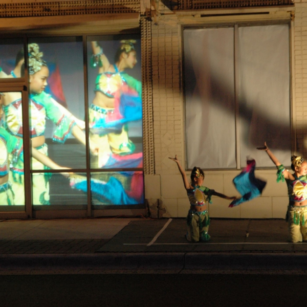 Evening, two dancers on a street with more dancers projected behind them.